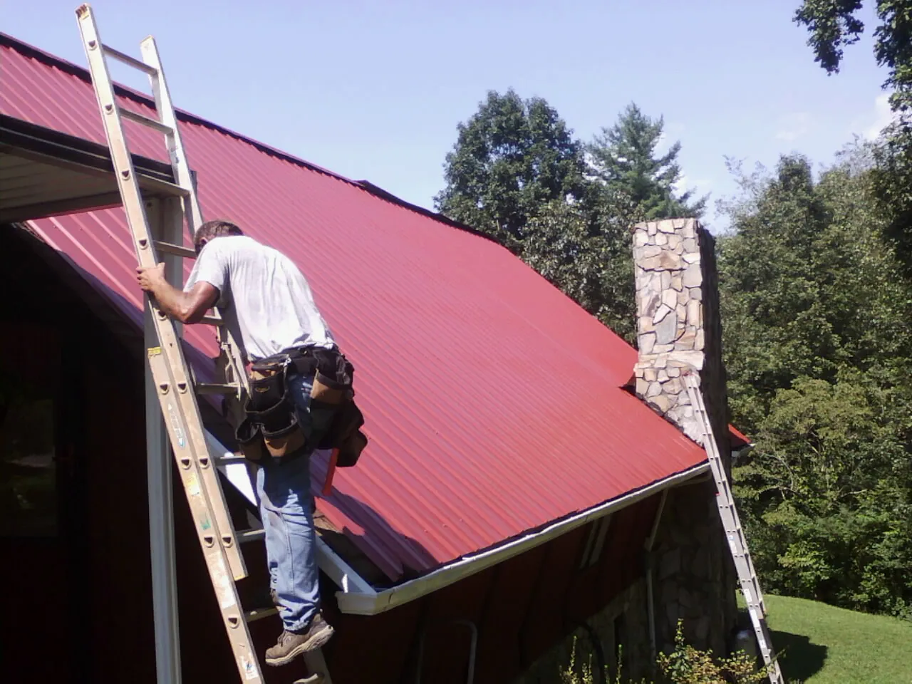 Roofer on a ladder beside a red metal roof and stone chimney — Soffit & Fascia Repair by Roofing Brothers in Furlong